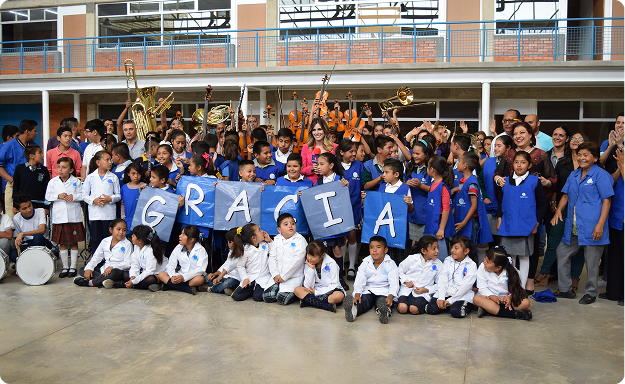 Grupo numeroso de niños y niñas en uniforme escolar posando junto a maestros y personal en un patio escolar. Los niños del centro sostienen carteles azules que forman la palabra ‘GRACIAS’, mientras detrás se observa una banda musical con instrumentos de metal y cuerdas levantados en señal de celebración.